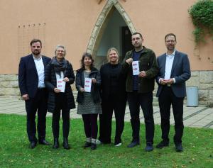 Drei Frauen und drei Männer stehen nebeneinander vor dem Verwaltungsgebäude Am Anger 15, drei halten einen Flyer in der Hand.