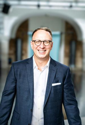 Ein Mann mit weißem Hemd und blauem Sakko steht vor einem Gebäude