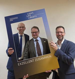 Drei Männer halten einen Aufsteller in Form eines Polaroid-Bildes und schauen hindurch. Auf dem Schild steht "Exzellent zusammen"
