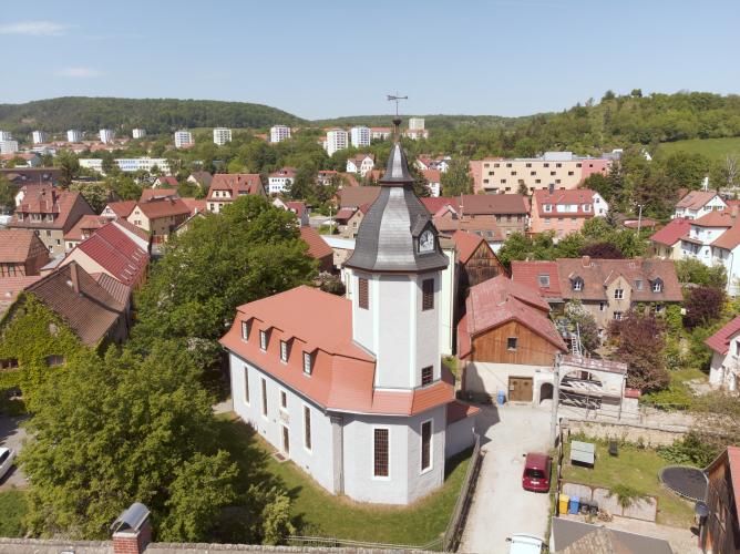 Eine Kirche mit kleinen Häusern, im Hintergrund moderne Neubauten und ein Bergkamm