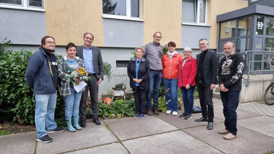 Neun Personen stehen vor einem gepflegten Blumenbeet vor einem Wohnblock