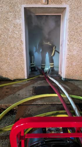 Ein Feuerwehrmann steht im Eingang eines verrauchten Hauses, Schläuche führen ins Innere des Gebäudes.