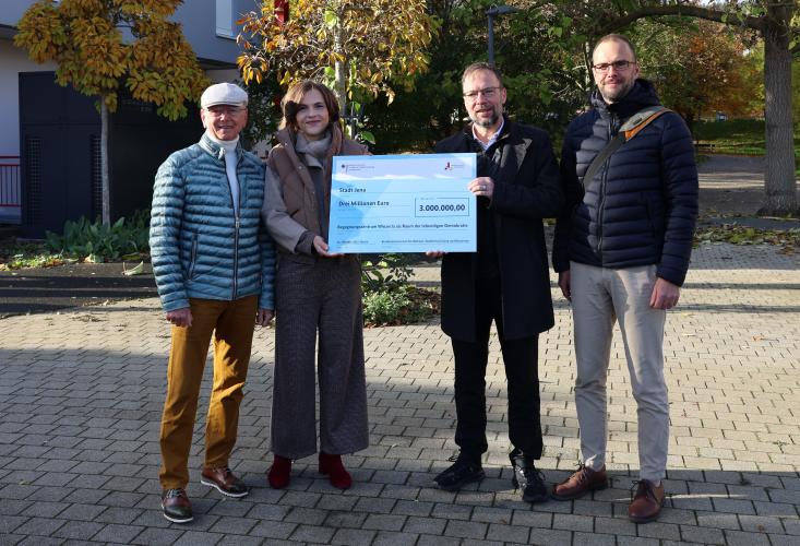 v.l.: Friedrich Wilhelm Gebhardt, ehemaliger Ortsteilbürgermeister von Jena-Winzerla, Ostbeauftragte Elisabeth Kaiser, Oberbürgermeister Thomas Nitzsche und Lars Liebe, Fachdienstleiter Stadtentwicklung stehen nebeneinander und halten einen großen Scheck.