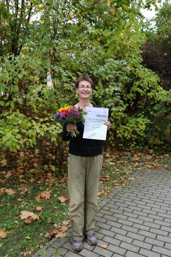 Eine Frau steht vor einer Grünfläche und hält einen Blumenstrauß und eine Urkunde in der Hand.