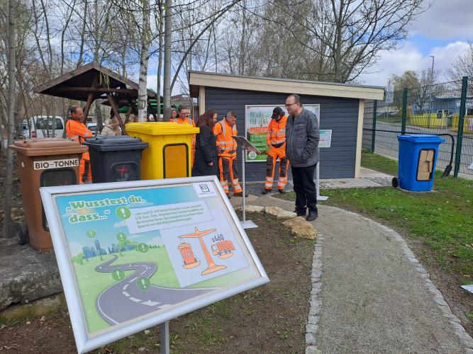 Vor einem kleinen Haus stehen ein Schild und drei Schau-Mülltonnen sowie mehrere Personen. 