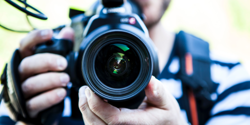 Ein Mann hält eine Fotokamera in der Hand, der Betrachter des Bildes sieht direkt in das Objektiv