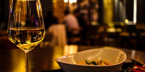 Ein Weinglas und ein Teller mit Essen auf einem Tisch in einem angenehm dunklen Raum, im Hintergrund sitzen Mneschen an einem Tisch, die unscharf zu sehen sind