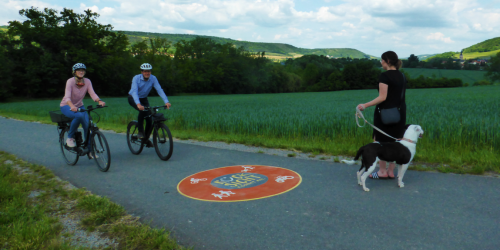 Drei Menschen auf einem Fahrradweg, zwei mit Fahrrad, eine Dame mit Hund
