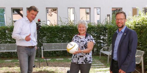 Drei Mensch schauen in die Kamera, die Frau in der Mitte hält einen Fußball.