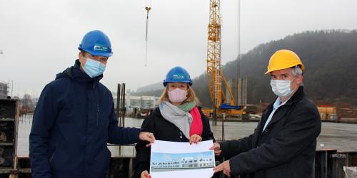 Drei Personen halten einen Bauplan in die Kamera. Sie stehen auf einer Baustelle.