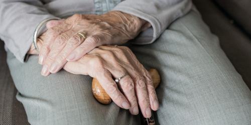 Blick auf Hände und Beine einer älteren sitzenden Person die einen Stock in der einen Hand hält