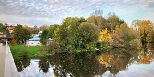 Im Vordergrund ein Fluss mit einer Brücker und im Hintergrund ein Zirkuszelt im Grünen