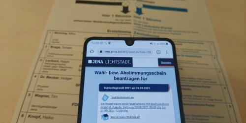 Ein Wahlschein liegt auf einem Tisch, darüber ein Smartfone mit der geöffneten Seite um die Briefwahl zu beantragen.