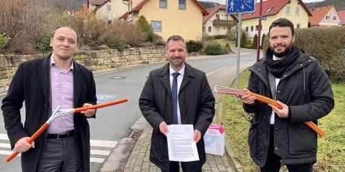 Drei Männer scheuen in die Kamera, im Hintergrund eine Siedlung mit Einfamilienhäusern