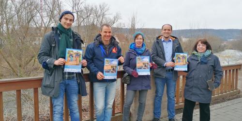 5 Menschen halten Plakate in die Kamera, hinter ihnen ein Fluss