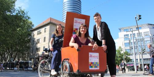 Drei Frauen. Eine sitzt auf einem Lasetnfahrrad, eine sitzt in dem Gepäckteil und eine steht daneben.
