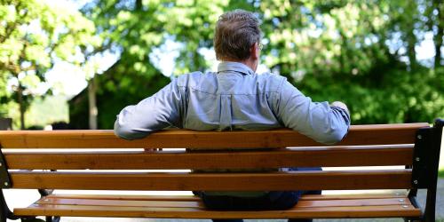 Ein älterer Mann sitzt auf einer Parkbank. Man sieht ihn von hinten.