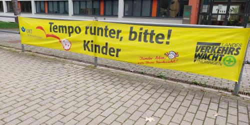 Vor einer Schule hängt ein Banner, dass zu Vorsicht im Verkehr aufruft.