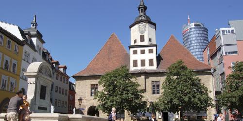 Im Vordergrund ein steinerner Brunnen auf einem großen Platz, im Hintergund mehrere Gebäude.