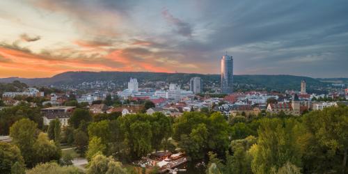 Panoramaaufnahme der Stadt Jena mit einem Fluss und viel Grün runsherum im Vordergrund und Bebauung im Hintergrund