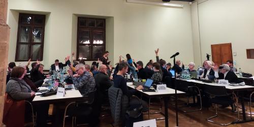 Stadtratsmitglieder sitzen an Tischen im Historischen Rathaus und heben die Hände, weil sie für einen Beschluss stimmen