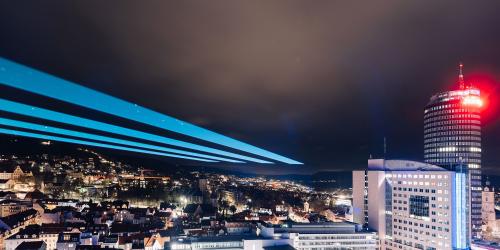 Das Foto zeigt Jena im Dunkeln mit erleuchteten Gebäuden und einem großen Laserstrahl am Himmel.