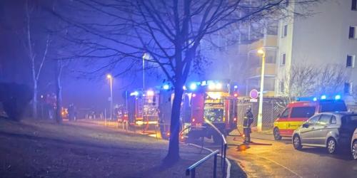 Blick auf eine Straße mit Plattenbauten. Davor stehen Feuerwehrautos mit Blaulicht