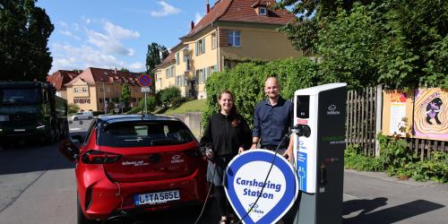 Eine Frau und ein Mann stehen zwischen einem Auto und einer E-Ladesäule. Die Frau hält ein Ladekabel in der Hand und der Mann ein Schild mit der Aufschrift: Carsharing Standort teilAuto.