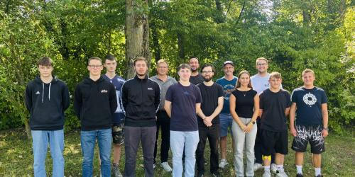 14 Personen stehen für ein Gruppenbild in zwei Reihen neben- bzs. hintereinander