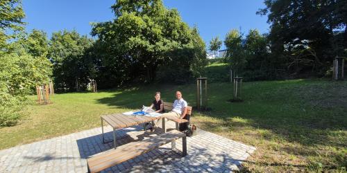 Naturnahe Lichtung mit Rastplatz: Eine Frau und ein Mann sitzen in der Sonne auf einer Bank.