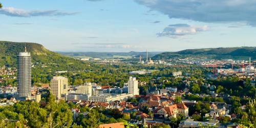 Das Foto zeigt eine weite Panoramaaufnahme der Stadt Jena in Thüringen, aufgenommen vom Landgrafenblick aus. Im Vordergrund sind grüne Baumkronen zu sehen, dahinter breitet sich die Stadt mit ihren Wohnhäusern und Bürogebäuden aus. Links ragt der runde Jentower deutlich über die übrigen Gebäude hinaus. Im Hintergrund ziehen sich die bewaldeten Hügel des Saaletals entlang, und der Himmel ist leicht bewölkt mit Sonnenlicht, das die Landschaft in warmes, freundliches Licht taucht.