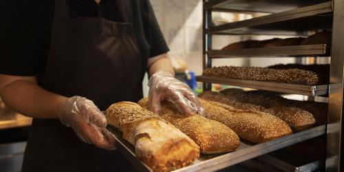 Eine Person in dunkler Arbeitskleidung und transparenten Einweghandschuhen hält ein Blech mit frisch gebackenen, langen Broten, teilweise mit Sesam bestreut. Im Hintergrund steht ein Metallregal mit weiteren Broten in einer Backstube.