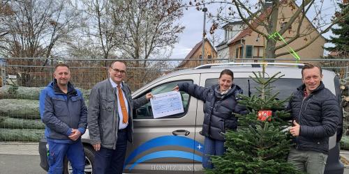 drei Männer und eine Frau stehen mit einem Spendenscheck und einem Weihnachtsbaum vor einem Auto.