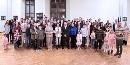 Große Gruppe von Menschen unterschiedlichen Alters steht gemeinsam in einem hellen Saal mit Parkettboden und hohen Fenstern für ein Gruppenfoto.