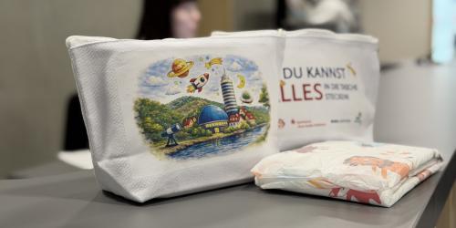 Zwei weiße Stofftaschen mit buntem Aufdruck stehen auf einem Tisch, daneben liegen Windeln; im Hintergrund unscharf Personen in einem Innenraum.