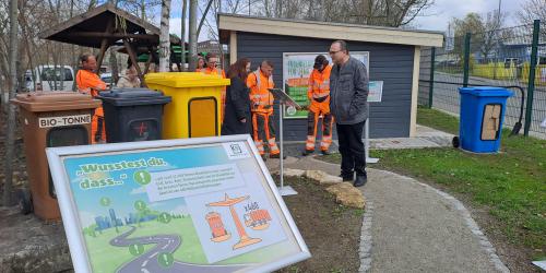 Vor einem kleinen Haus stehen ein Schild und drei Schau-Mülltonnen sowie mehrere Personen. 
