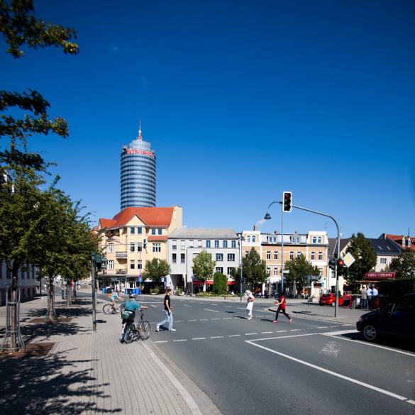 Menschen überqueren eine Straße an einer Ampelkreuzung, im Hintergrund ein großer Turm.