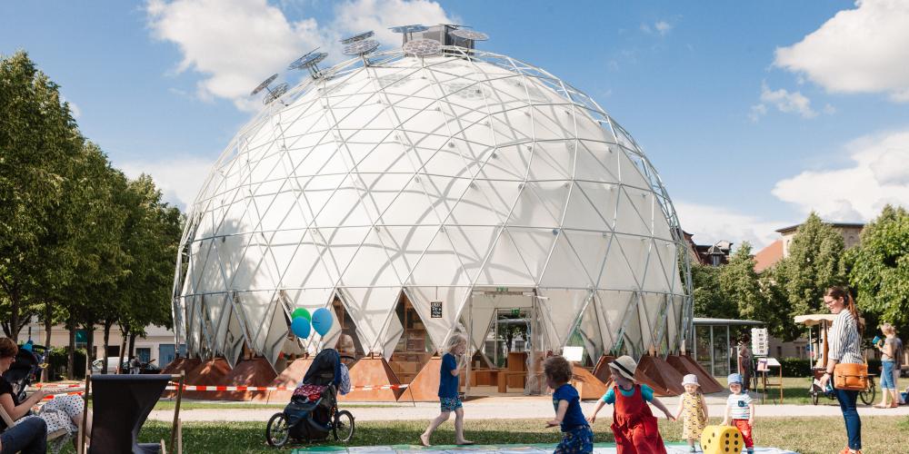 Kinder spielen in einem Park vor einem kuppelförmigen Pavillion.