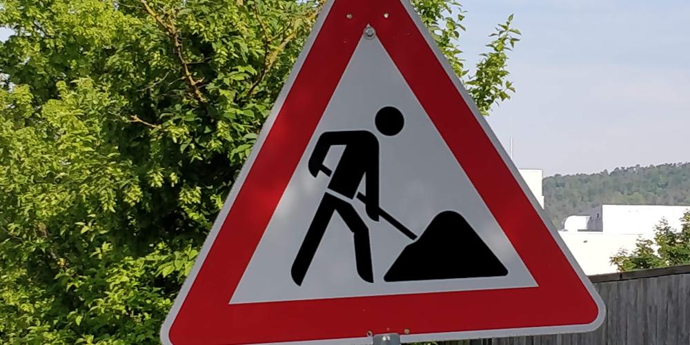 Ein Baustellenschild steht vor einem Baum am Straßenrand.