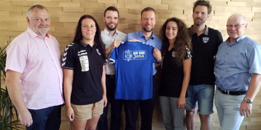 Fünd Männer und zwei Frauen halten ein Fußball-Trikot in die Kamera.