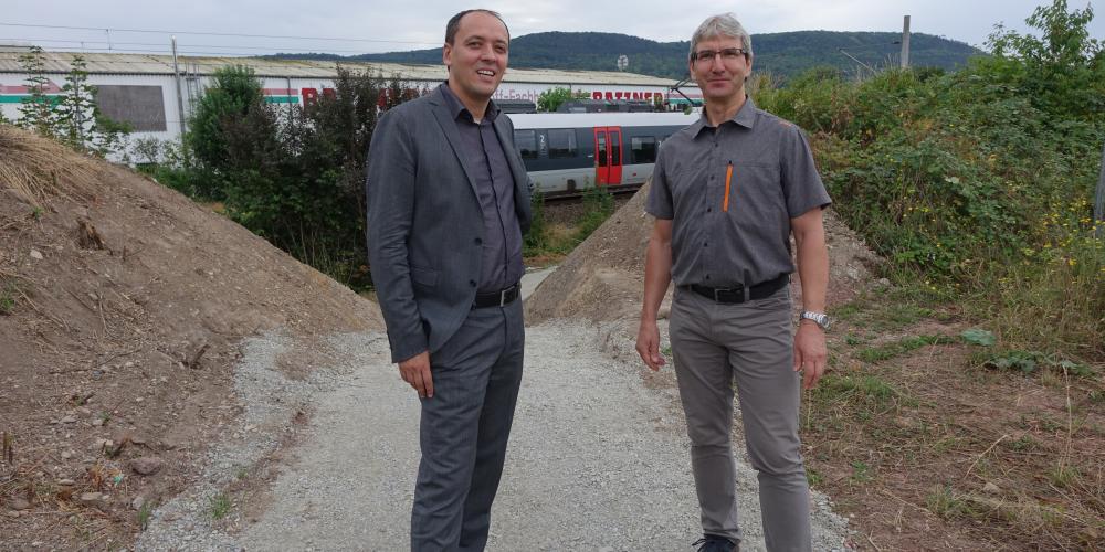 Bürgermeister Christian Gerlitz und KSJ-Mitarbeiter Jürgen Henning stehen auf einem geschotterten Weg. Der Weg führt durch einen Einschnitt in einem Lärmschutzwall. Im Hintergrund fährt ein Zug vorbei.