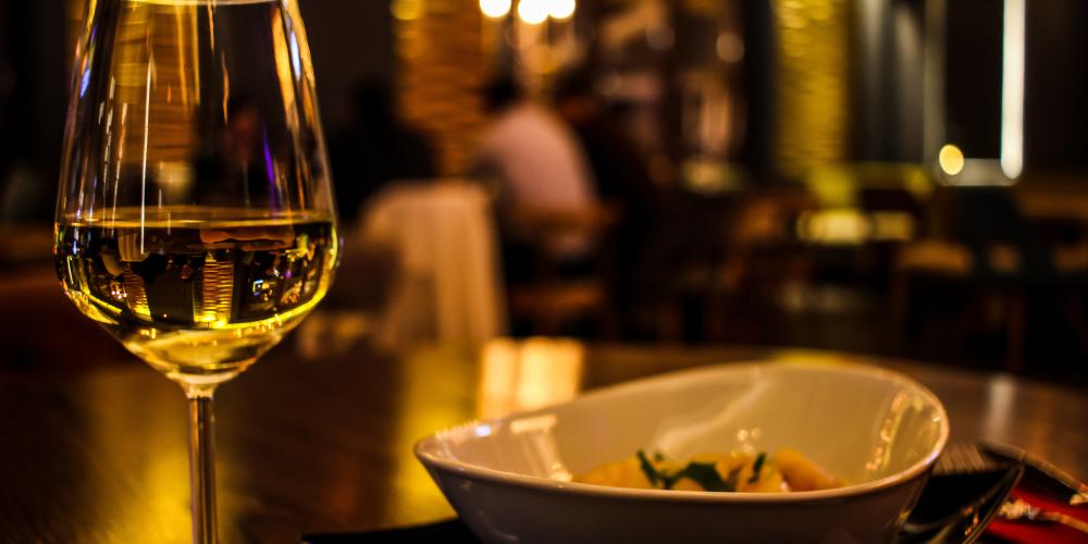 Ein Weinglas und ein Teller mit Essen auf einem Tisch in einem angenehm dunklen Raum, im Hintergrund sitzen Mneschen an einem Tisch, die unscharf zu sehen sind