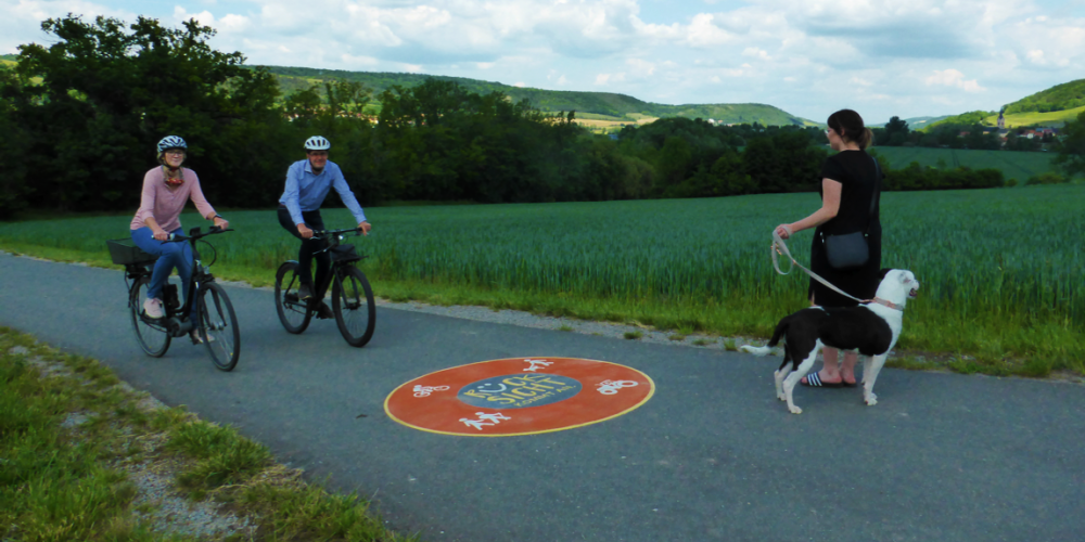 Drei Menschen auf einem Fahrradweg, zwei mit Fahrrad, eine Dame mit Hund