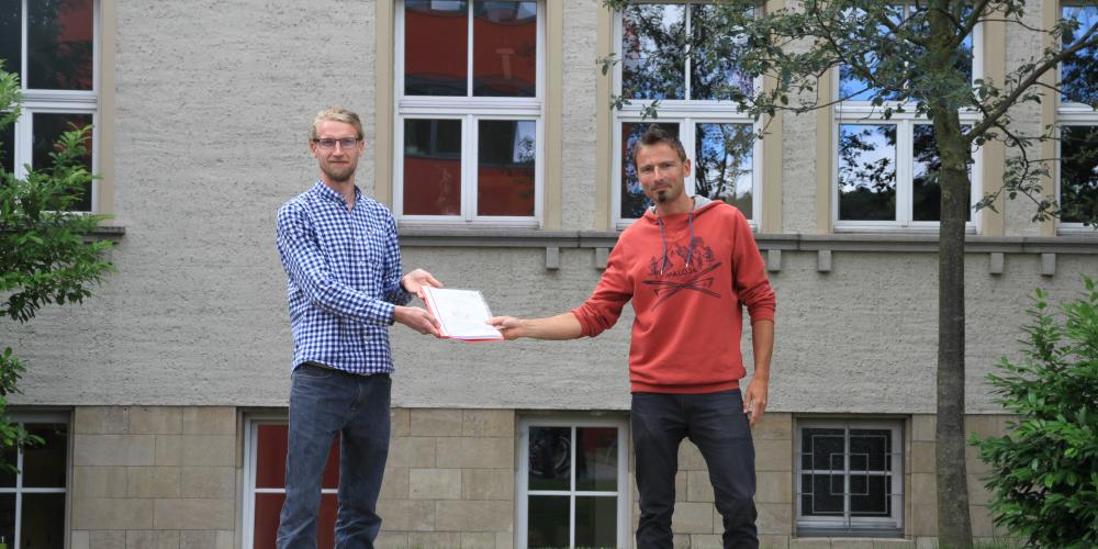 Zwei Personen stehen vor einem Gebäude. Die eine Person übereicht ein Dokument der anderen.