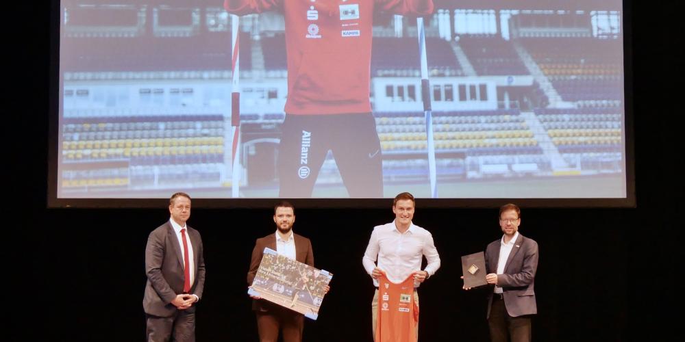 Vier Männer auf ein Bühne, im Hintergrund ein großes Bild eines Sportlers