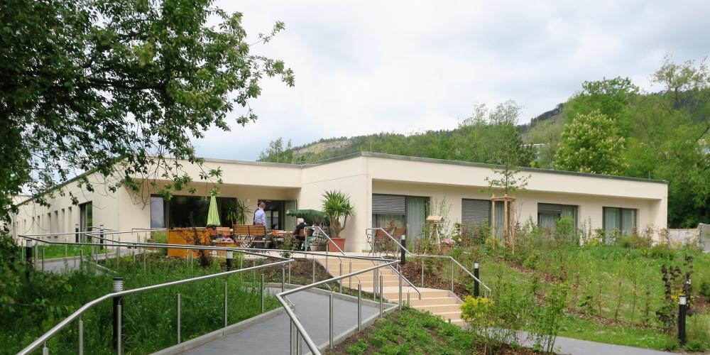 Blick auf eine flaches helles Gebäude, im Vordergrund eine Treppe und eine Rampe die durch einen Garten zum Haus führt.