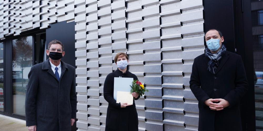 Zwei Herrn und eine Dame schauen in die Kamera. Im Hintergrund eine Fassade aus Metall.