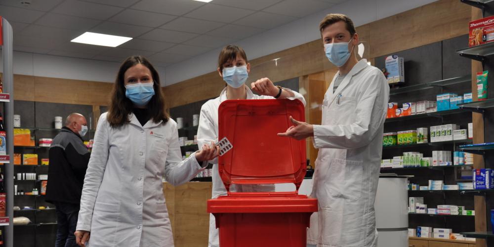 Drei Menschen stehen vor einer Box, in welche sie Medikamentenverpackungen werfen. Im Hintergrund befinden sich viele Regale.