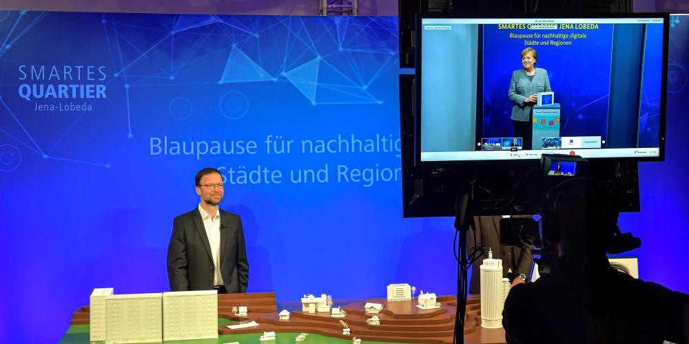 Ein Mann steht in einem Filmaufnahmestudio vor einem blauen Hintergrund. Rechts neben ihm sieht ist auf einem eine Frau zu sehen. Das Bild ist aus Perspektive des Technikpults aufgenommen worden.