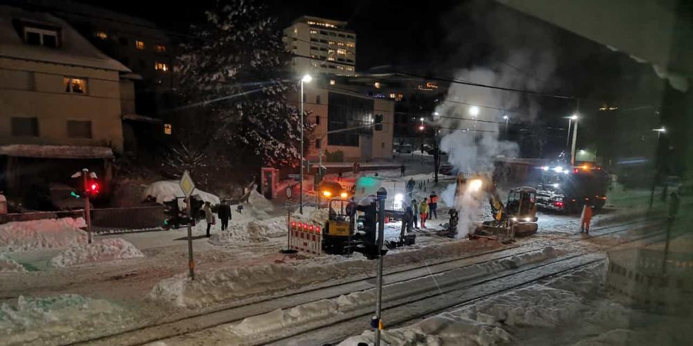 Blick auf die Kreuzung bzw. verschneite Straße im Dunkeln, zu sehen sind Arbeiter und Maschinen